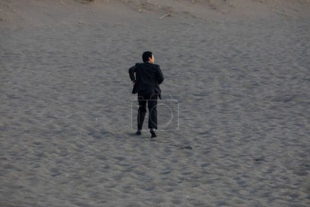 Photo for Asian businessman in formal suit walking along the sandy beach, rear view - Royalty Free Image