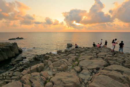 Téléchargez les photos : Touristes debout sur des rochers et profitant d'un magnifique coucher de soleil sur la mer - en image libre de droit