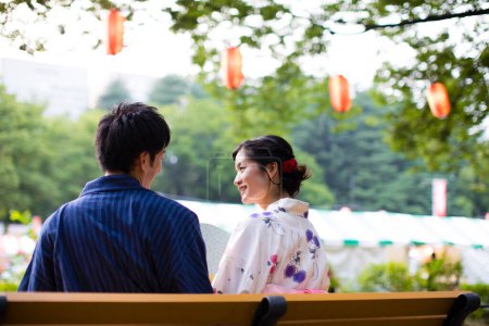 Photo for Back view of young Japanese couple wearing traditional kimono sitting on bench in summer park - Royalty Free Image
