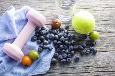 Foto de Tomates, arándanos, vaso de agua, toalla, pelota de tenis y mancuerna sobre mesa de madera - Imagen libre de derechos