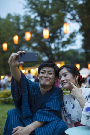 Photo for Young Japanese couple wearing traditional kimono taking selfie on smartphone in evening park - Royalty Free Image