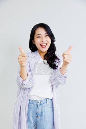 Foto de Feliz asiático mujer en camisa y jeans posando con los pulgares hacia arriba gesto aislado en blanco fondo - Imagen libre de derechos