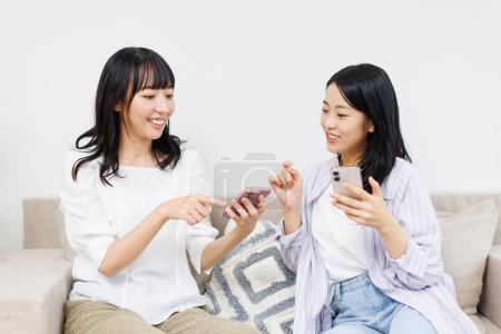 Photo for Two asian women sitting on couch at home, talking and using smartphones - Royalty Free Image