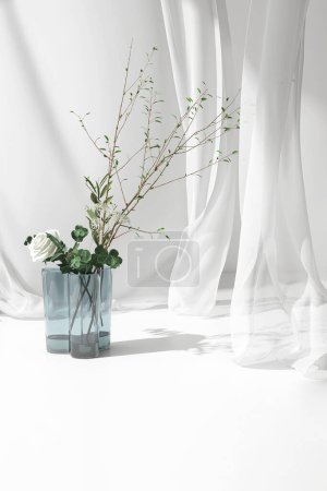 Foto de Ramo de rosas blancas y rama de árbol verde en jarrón de vidrio azul en la mesa de mostrador, suave soplado cortina de tela pura a la luz del sol para cosméticos de lujo, cuidado de la piel, belleza, pantalla de productos de moda de fondo 3D - Imagen libre de derechos