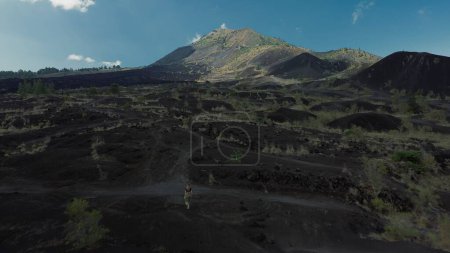 Photo for Top view of a young woman who walks towards Batur volcano, Bali island, on black lava. A beautiful view of the mountain, the consequences of a volcanic eruption in the form of black lava. - Royalty Free Image