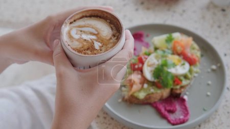 Photo for Morning freshly brewed coffee in a cute coffee shop, coffee, and avocado toast for breakfast. Close shot of female hands holding a cup of coffee against the background of toast for breakfast. - Royalty Free Image