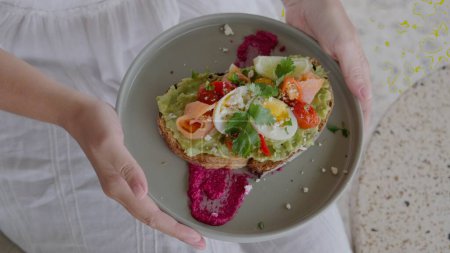 Photo for Healthy breakfast in a cafe, a girl holds a plate with avocado toast, with the addition of smoked salmon, cherry tomatoes, boiled egg, goat cheese and beetroot hummus. The concept of proper nutrition. - Royalty Free Image