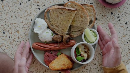 Téléchargez les photos : Petit déjeuner traditionnel britannique dans une assiette prise par un homme. Un gros plan de mains masculines prenant une assiette de petit déjeuner traditionnel qui comprend des œufs bénédicte, saucisses frites, bacon et pain grillé. - en image libre de droit