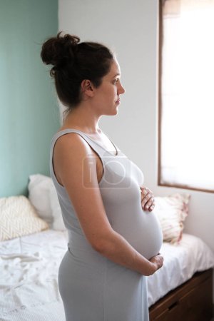 Photo for Medium shot side view of a pregnant brunette woman with lush hair gathered in a bun in a light dress. A pregnant woman wraps her arms around her stomach, looking out the window and smiles. Calm. - Royalty Free Image