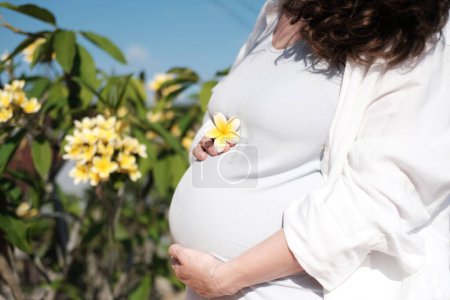 Foto de Una joven embarazada se encuentra en un balcón con vistas a un jardín con un árbol con flores y acaricia su vientre. Acariciando la barriga en la que el bebé está esperando nacer. Feliz maternidad.. - Imagen libre de derechos