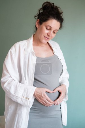 Foto de Foto vertical de la futura madre. Una mujer embarazada con un gran vientre en su tercer trimestre dobló sus manos en forma de corazón sobre su barriga. Esperando un bebé y padres felices. - Imagen libre de derechos