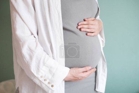 Photo for Side view of a pregnant woman in a light shirt and with a big belly. The woman hands her arms around her pregnant belly. Waiting for a baby, happy moments of motherhood. - Royalty Free Image