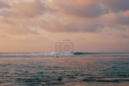 Foto de Silueta irreconocible de un surfista masculino con una tabla de surf en el océano al atardecer. El cielo está pintado en tonos de rosa, el océano refleja el cielo, un surfista camina hacia. - Imagen libre de derechos