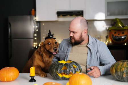 Photo for Front view of a man sitting at a kitchen table with a golden retriever dog and carving a pumpkin for a Halloween party. Preparing for a Halloween party, DIY decorations. - Royalty Free Image