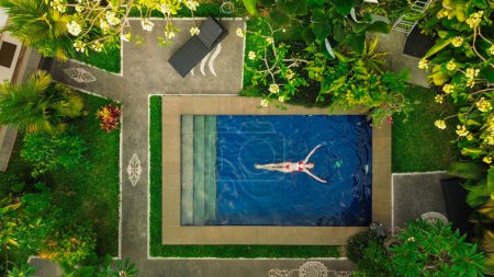 Photo for Birds eye view of a blue water swimming pool located in the jungle on a tropical. A young beautiful woman in a red swimsuit lies in the pool. Vacation and travel concept to tropical islands. - Royalty Free Image