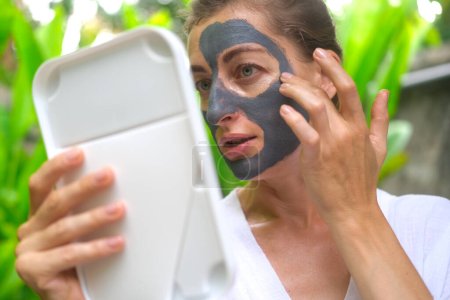 Téléchargez les photos : Une femme d'âge moyen applique un masque d'argile grise sur son visage et se regarde dans le miroir. Une femme dans une cour sur fond de feuillage vert sous les tropiques applique un masque d'argile pour nettoyer sa peau. - en image libre de droit