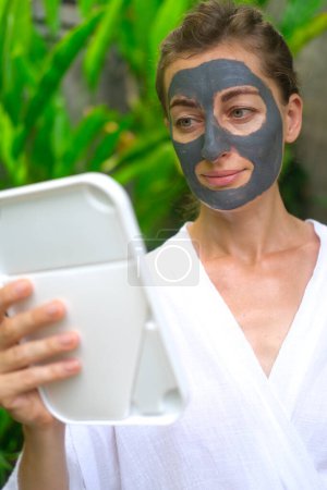Photo for Vertical shot a woman applies a gray clay mask to her face and looks in the mirror. A woman in a courtyard against a backdrop of green foliage in the tropics applies a clay mask to cleanse her skin. - Royalty Free Image