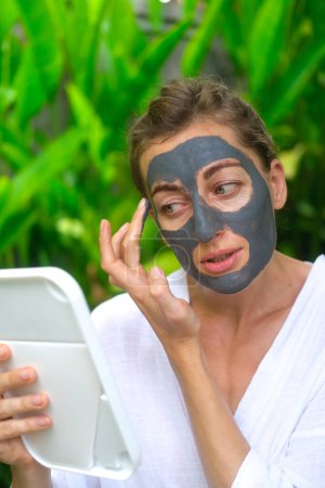 Photo for Vertical shot a woman applies a gray clay mask to her face and looks in the mirror. A woman in a courtyard against a backdrop of green foliage in the tropics applies a clay mask to cleanse her skin. - Royalty Free Image