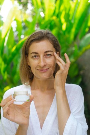 Photo for Vertical close up photo of the face of a middle-aged woman. A woman applies cream under her eyes with massage movements and holding a jar in her hand. Anti aging skin care. - Royalty Free Image