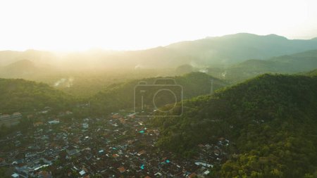 Foto de Una pintoresca vista aérea del pueblo de Padang Bai cerca del puerto en la isla de Bali. El pueblo está rodeado de montañas y colinas verdes. Atardecer dorado, el sol se pone en las montañas. - Imagen libre de derechos
