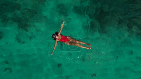 Foto de Vista aérea de una joven en traje de baño rojo que nada con una máscara cerca de la orilla en el océano y observa el arrecife de coral. Buceo en el océano, agua de color esmeralda. - Imagen libre de derechos
