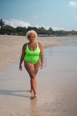 Foto de Foto vertical, una mujer afroamericana de cuerpo entero y talla grande con el pelo rizado en un traje de baño verde claro camina a lo largo de la playa cerca del océano. El concepto de aceptación corporal y positividad corporal. - Imagen libre de derechos