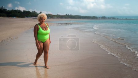 Téléchargez les photos : Une femme afro-américaine pleine longueur, plus la taille avec les cheveux bouclés dans un maillot de bain vert clair se promène le long de la plage près de l'océan. Le concept d'acceptation corporelle et de positivité corporelle. Bannière, espace libre. - en image libre de droit
