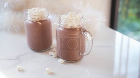 Photo for Two transparent mugs with cold cocoa with marshmallows stand on a white kitchen table. There is Christmas tinsel in the background. Christmas summer background image with refreshing drink. - Royalty Free Image