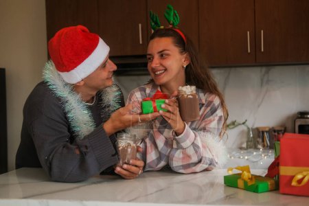 Foto de Familia en la cocina en Navidad, un hombre con un sombrero de Santa le da a su esposa en una diadema de Navidad con cuernos de ciervo un regalo. Felicidad de recibir regalos para Navidad y Año Nuevo. - Imagen libre de derechos