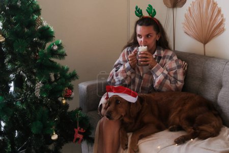 Foto de Una joven vestida con un traje navideño con cuernos de reno se sienta en el sofá y bebe cacao con malvaviscos. Un perro recuperador de oro en un sombrero de Santa se encuentra junto a ella. Acogedora Navidad con un perro. - Imagen libre de derechos