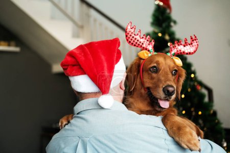 Foto de Vista posterior de un hombre con un sombrero de Santa Claus abrazando a un perro recuperador de oro con una diadema con astas de ciervo contra el telón de fondo de un árbol de Navidad con decoraciones y guirnaldas. Amantes del perro. - Imagen libre de derechos