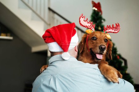Foto de Abrazos de un hombre en un sombrero de Santa con un perro en un sombrero con astas de ciervo sobre el fondo de un árbol de Navidad con guirnaldas y decoraciones. Feliz Año Nuevo y Feliz Navidad. Postal para una tienda de mascotas - Imagen libre de derechos
