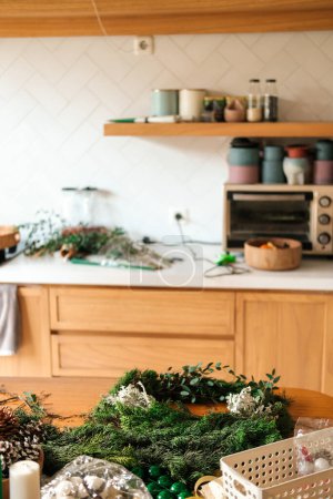 Foto de Ambiente navideño en la cocina. Sobre la mesa de madera hay unas ramas de abeto, de que es hecha la corona. También hay velas en la mesa, piñas y bolas para el árbol de Navidad. - Imagen libre de derechos