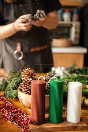 Photo for Small business producing Christmas wreaths and decorations. The hands of a young woman create a wreath of fir branches and decorations in the form of berries and cones. DIY holiday decor. - Royalty Free Image