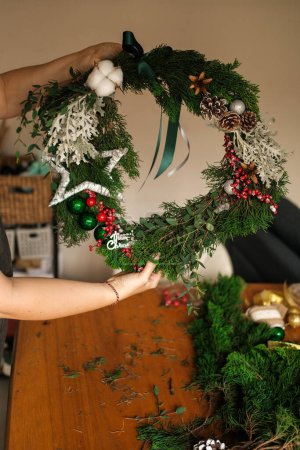 Photo for Small business producing Christmas wreaths and decorations. The hands of a young woman create a wreath of fir branches and decorations in the form of berries and cones. DIY holiday decor. - Royalty Free Image