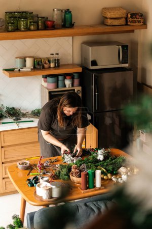 Téléchargez les photos : Une jeune femme à la maison dans sa cuisine se prépare pour la célébration de Noël et fait une couronne de branches de sapin sur une table en bois. Atmosphère familiale de préparation pour le Nouvel An. - en image libre de droit