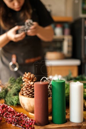 Photo for In the foreground are wax candles in Christmas colors. In the background, a young woman creates a Christmas wreath from fir branches in her kitchen. Preparations for the main winter holiday. - Royalty Free Image