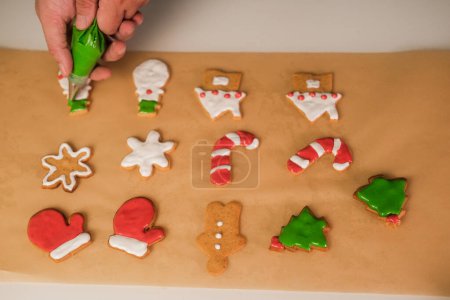 Téléchargez les photos : Les mains de l'homme tiennent un sac à pâtisserie contenant du glaçage vert. Il fait des biscuits au pain d'épice pour Noël. Cookies en forme de flocon de neige, bonhomme de neige et sapin de Noël. - en image libre de droit