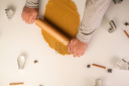 Photo for Top view of a man hands rolling out gingerbread dough for Christmas with a wooden rolling pin on a white table. Cookie cutters and spices are laid out on the table. - Royalty Free Image