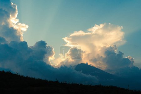 Téléchargez les photos : Vue sur le sommet du volcan Agung symbole de l'île de Bali, qui a disparu derrière les nuages. Volcan au coucher du soleil dans les nuages pelucheux, les rayons du soleil se brisent à travers les nuages. - en image libre de droit