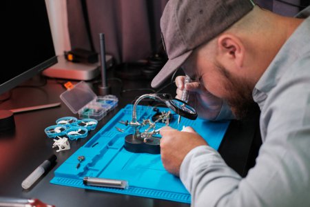 Foto de Fotografía media de un ingeniero masculino soldando el chip del transmisor de vídeo de un dron FPV con una lupa. Centro de servicio para la reparación y mantenimiento de equipos. Reparación de drones. - Imagen libre de derechos
