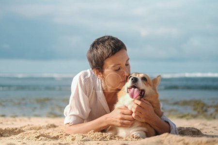 Photo for Portrait of a woman with dark hair and a short haircut kissing her Welsh Corgi dog while walking on the beach. Love and affection for your pets. Woman and corgi on the beach. - Royalty Free Image