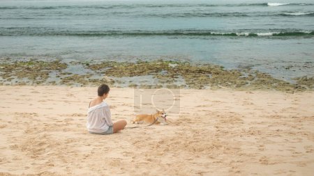 Téléchargez les photos : Vue arrière d'une femme en chemise blanche assise sur la plage avec un Corgi gallois en laisse. Le chien est fatigué et s'allonge sur le sable avec sa langue suspendue. Promenade chien concept de service. - en image libre de droit
