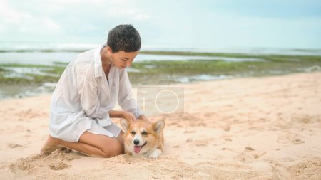 Photo for A middle-aged woman in a white shirt with a short haircut strokes her Welsh Corgi on the beach. The dog lies on the sand with its tongue hanging out. Traveling with a pet. Banner pet store. - Royalty Free Image