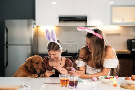 Téléchargez les photos : Maman et fille portant des porte-cheveux d'oreille de lapin s'assoient à la table de cuisine et décorent les œufs de Pâques. À côté d'eux se trouve un chien récupérateur d'or avec sa langue suspendue. Traditions familiales. - en image libre de droit