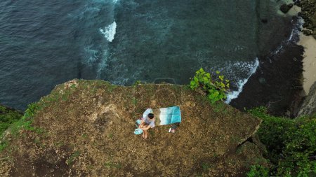 Photo for Top down view of a woman artist sitting on the edge of a cliff near the ocean and painting a picture on canvas. She paints a seascape. The process of drawing on the seashore as seen from a drone. - Royalty Free Image