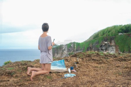 Téléchargez les photos : Une femme artiste est assise sur le bord d'une falaise et peint une image de la mer avec des peintures acryliques sur une toile couchée sur le sol. Concept d'inspiration nature et océan. Dessiner en plein air en été. - en image libre de droit