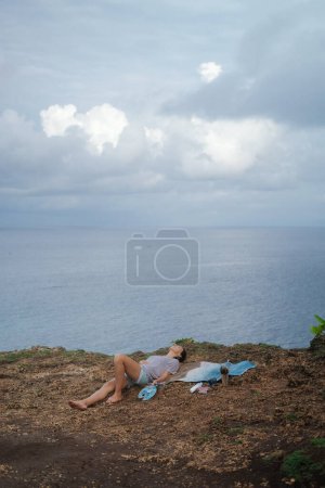 Téléchargez les photos : Une femme artiste se trouve sur le bord d'une falaise à côté d'une peinture qu'elle a peinte avec des peintures acryliques sur toile. Concept d'inspiration de la nature par l'océan. Dessiner à l'extérieur. - en image libre de droit