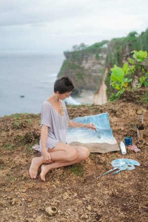 Foto de Una mujer artista se sienta en el borde de un acantilado y pinta un cuadro con pinturas acrílicas sobre un lienzo que yace en el suelo. Paisaje del mar al aire libre en verano. Dibujar un dibujo en la orilla del océano. - Imagen libre de derechos