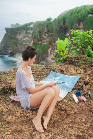Téléchargez les photos : Une femme artiste est assise sur une falaise au bord de l'océan et peint une peinture de paysage de la vie. Peinture de mer avec peintures acryliques sur toile. À côté de l'image se trouve une palette de peintures, un plein air en plein air. - en image libre de droit